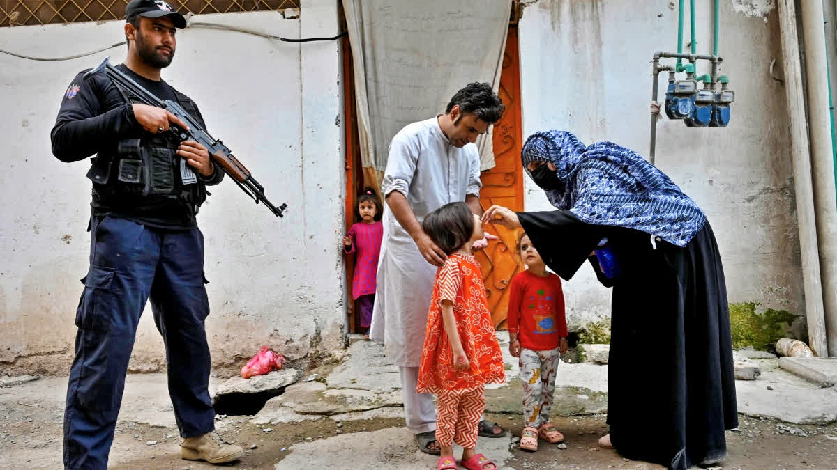 This photograph taken on October 29, 2024 shows an elite police personnel (L) standing guard as a health worker (R) administers polio drops to a child during a door-to-door poliovirus vaccination campaign on the outskirts of Peshawar.