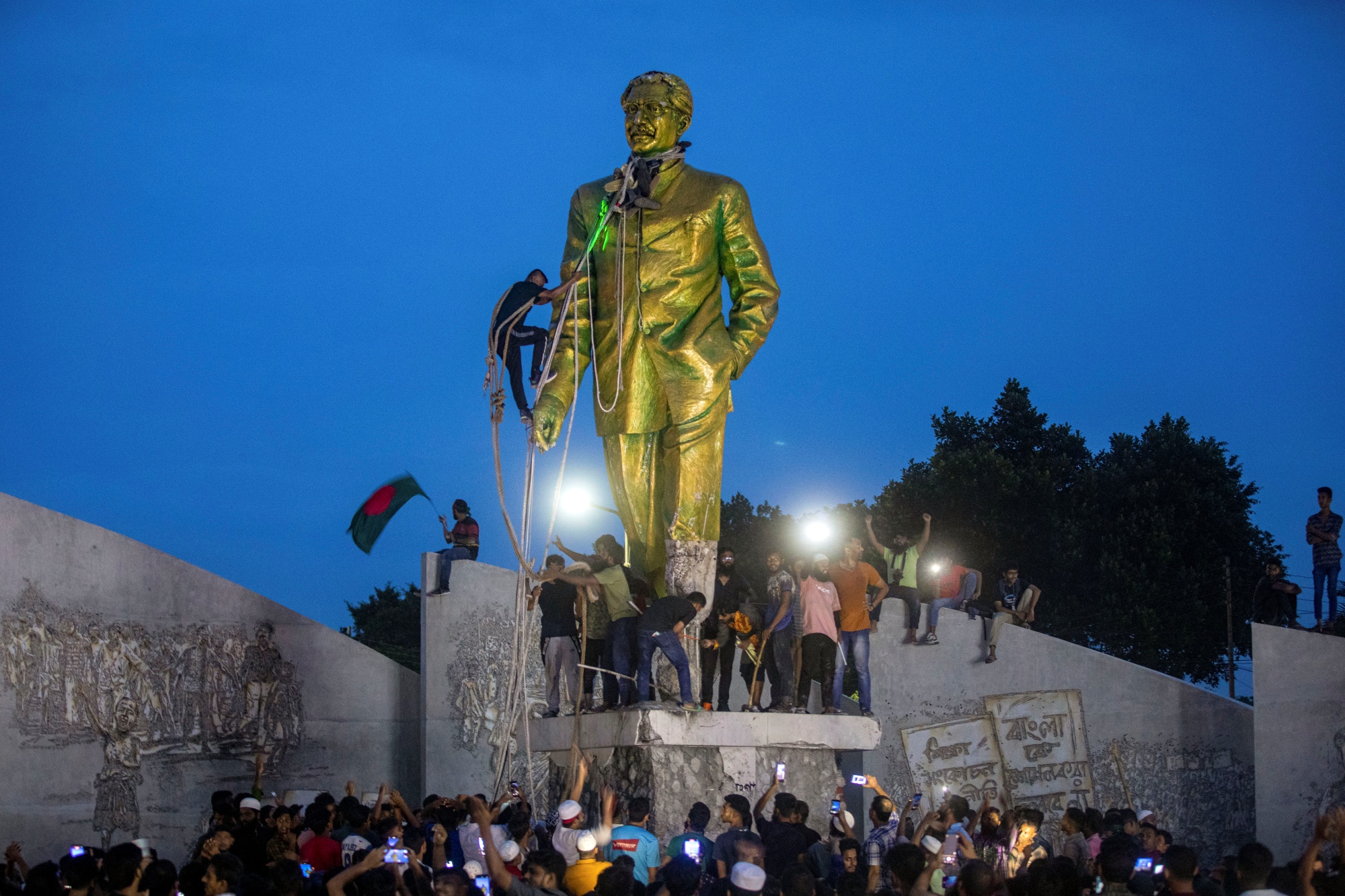 A man climbs to tie a rope around the head of a large statue of Sheikh Mujibur Rahman, father of Bangladesh leader Sheikh Hasina, as protesters try to bring it down after she resigned as Prime Minister, in Dhaka, Bangladesh, Monday, Aug. 5, 2024.