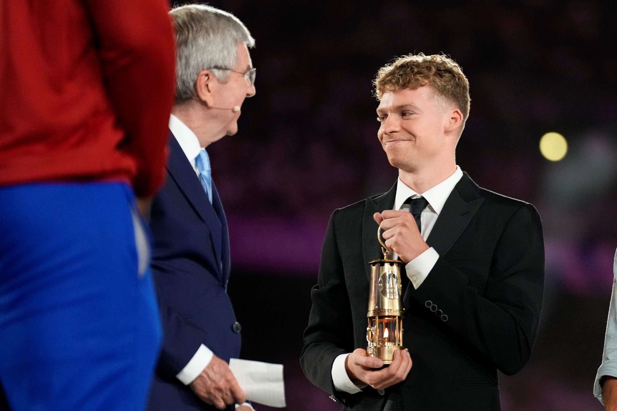 French swimmer Leon Marchand carries a lantern containing the Olympic flame with IOC President Thomas Bach, left, during the 2024 Summer Olympics closing ceremony at the Stade de France, Sunday, Aug. 11, 2024, in Saint-Denis, France.