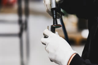 This image captures a technician, dressed in black and wearing white gloves, meticulously working on a small metallic object. The technician is using a tool that resembles an air ratchet, indicating the nature of work to be mechanical. The blurred objects in the background suggest an industrial or workshop setting.