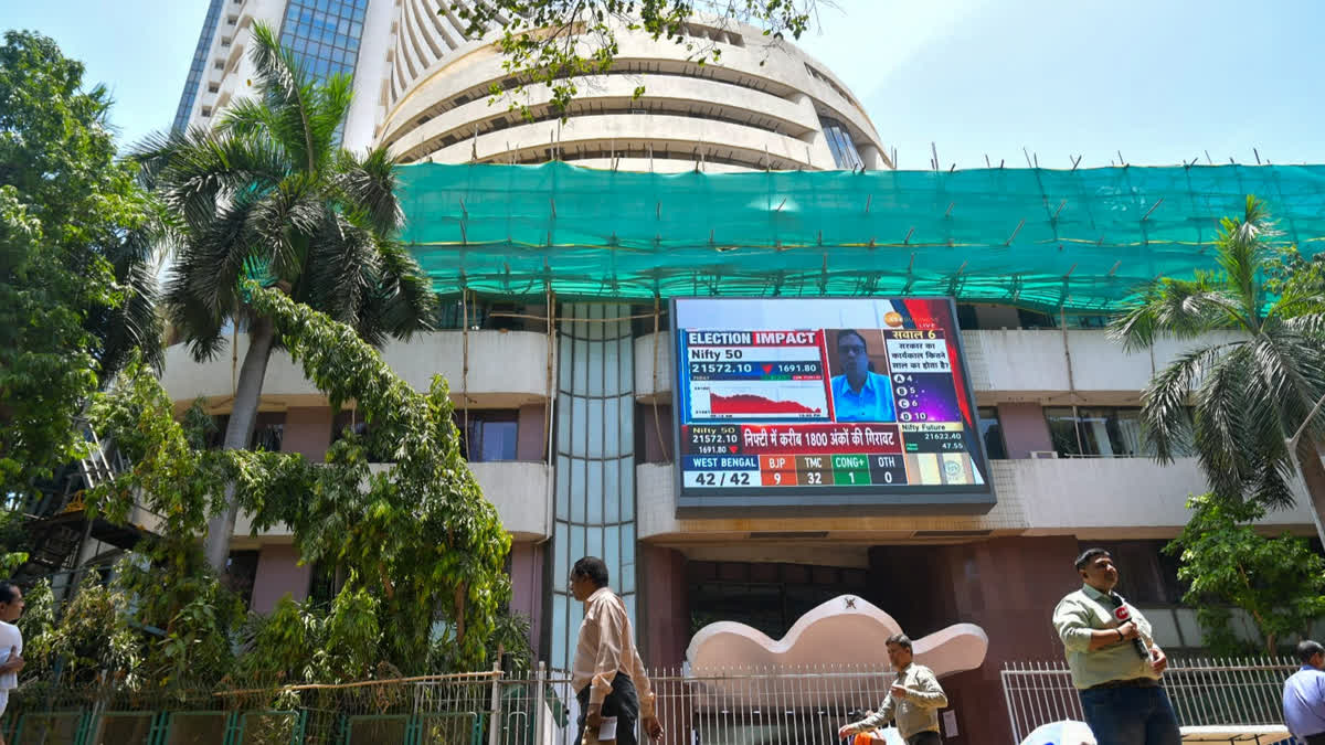 People walk past a screen showing stock market indices