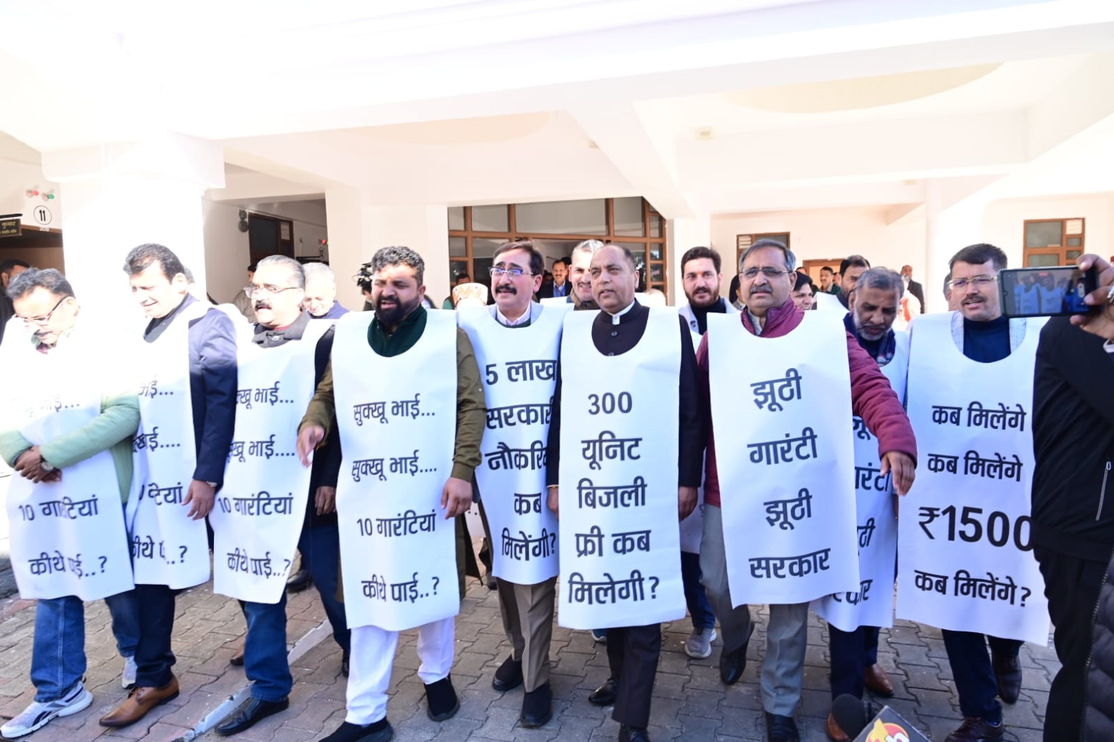 गले में कांग्रेस की गारंटियों के पोस्टर डालकर बीजेपी विधायकों का प्रदर्शन