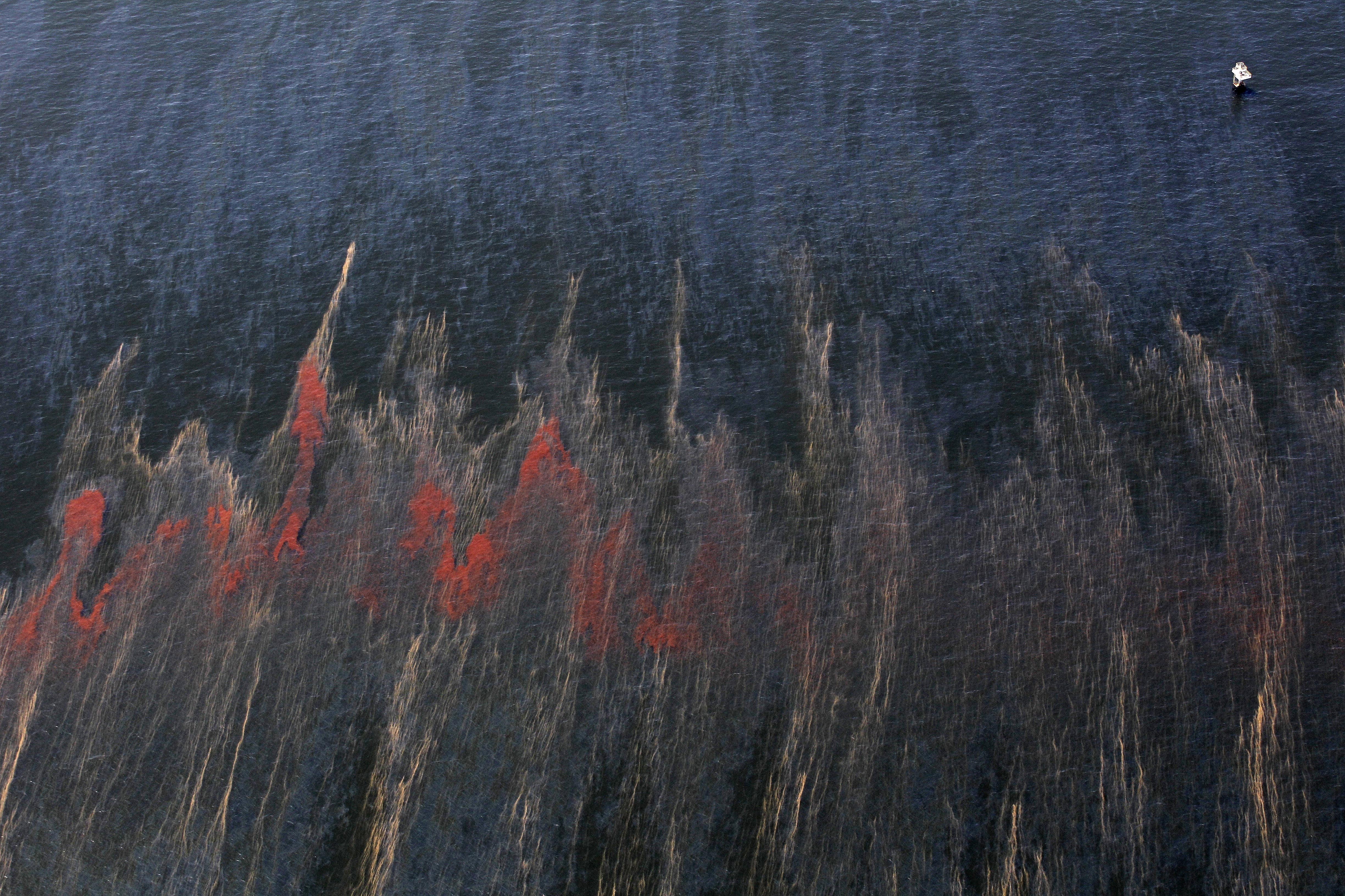 In this May 5, 2010 file photo, oil and oil sheen are seen moving past an oil rig, top right, in the waters of Chandeleur Sound, La. Ten years after an oil rig explosion killed 11 workers and unleashed an environmental nightmare in the Gulf of Mexico, companies are drilling into deeper and deeper waters where the payoffs can be huge but the risks are greater than ever.