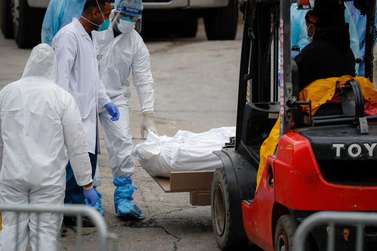 A body wrapped in plastic is prepared to be loaded onto a refrigerated container truck used as a temporary morgue by medical workers due to COVID-19 concerns